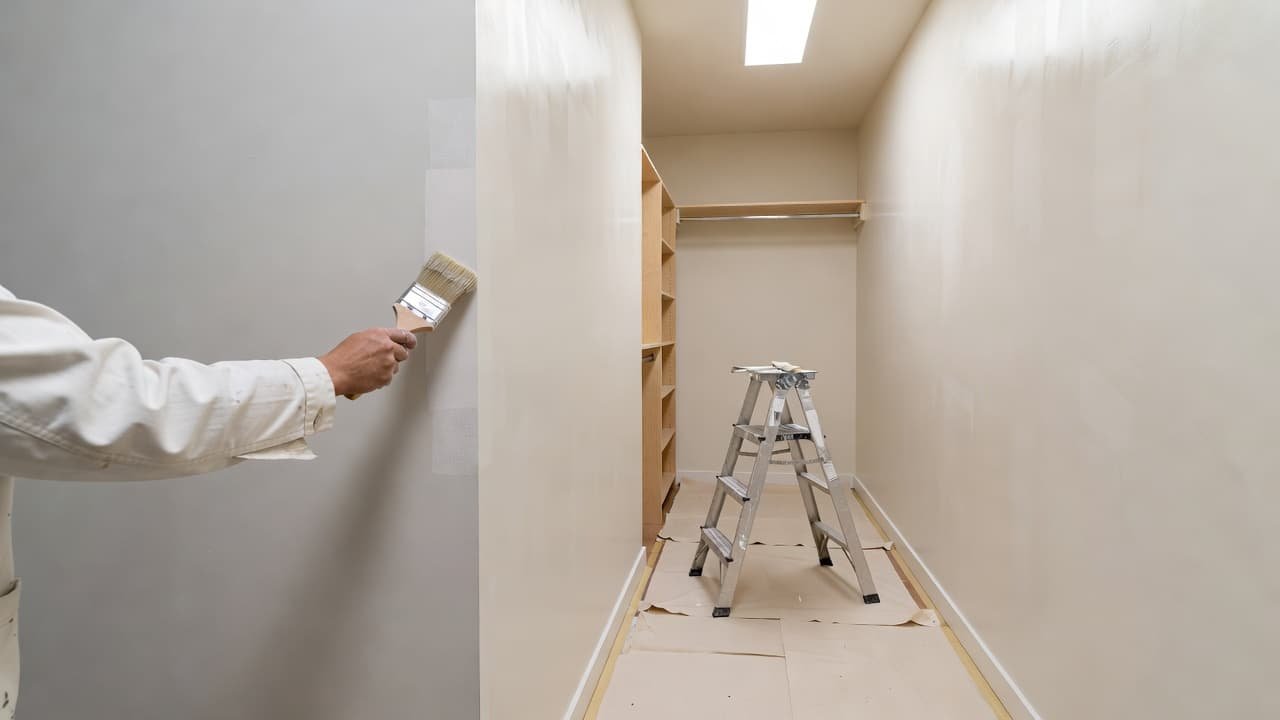 How Closet Painting Changes Whole-House Repaint Cost 2 A skilled painter in clean white work clothes uses a small brush to carefully apply paint to the corner of a walk-in closet wall under bright, even interior lighting conditions.